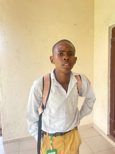 The media file shows a young Black male from the waist up, in what appears to be a school uniform. He is wearing a white collared shirt and khaki-colored shorts. The shirt is partially unbuttoned, revealing straps that go over his shoulders, possibly part of a backpack or a vest. The background is a plain, light-colored wall, with a darker trim at the top. The overall lighting suggests it might be daytime, possibly indoors or in a shaded outdoor area. The individual is looking directly at the camera with a slight smile. There are no overt indications of weather or a specific time of day beyond the general lighting. The location context provided is Unguwar Shanu, Nigeria, which is a neighborhood in Kano State. No text is visible in the image.