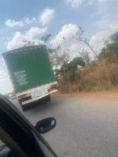 The image depicts a green box truck traveling on a paved road in Tataura, Nigeria, viewed from the driver's side of a following dark-colored vehicle. The truck's rear cargo area is green, featuring white and red reflective tape along its perimeter. A "DISCLAIMER" is printed on the back, stating, "THIS TRUCK IS FOR ANIMAL/VEGETABLES ONLY. ANY OTHER GOODS WILL BE CONFISCATED. WARNING! THIS IS ALL ABOUT DELIVERING! PLEASE CALL: 081260309067." The lower portion of the truck's rear is white. The road surface is gray asphalt with visible cracks, bordered by a brown dirt shoulder on the right. The environment to the right of the road consists of dry, brownish tall grasses and sparse, largely leafless trees. In the mid-ground, a small, light-colored building is visible amidst the vegetation, with a tall, thin structure partially obscured behind it. The sky is clear blue with scattered white cumulus clouds. The side-view mirror and a portion of the door frame of the observer's vehicle are visible in the lower foreground. No people are visible.