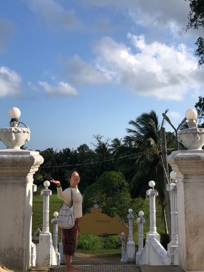 In the city of Pilana, Sri Lanka, a woman stands on a paved pathway, her body angled to the right, and her gaze directed upwards. Her right arm is extended upwards and outwards, with her palm facing upwards, as if to catch something from the sky. She wears a long-sleeved cream-colored shirt, burgundy shorts, and a white tote bag slung over her right shoulder. Her left hand is raised, with her palm also facing upwards. The background features lush green foliage, including palm trees, under a bright blue sky dotted with white clouds. White ornate pillars with spherical light fixtures flank the woman, framing the pathway and the tropical landscape.