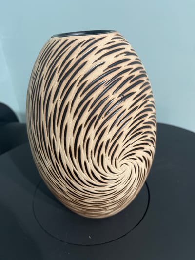 This is a close-up, eye-level, medium shot of a ceramic vase with a distinctive carved pattern. The vase is oval-shaped and its surface is decorated with black, curved lines that create a swirling, almost hypnotic effect, resembling a vortex or the texture of wood grain. The base material of the vase appears to be a light, earthy tone. It is positioned on a dark, circular object, likely a stand or part of a display. In the background, a wall is visible, half of which is a muted blue-grey and the other half a dark, possibly black, surface that blends with the object the vase is resting on. The lighting is bright and even, casting subtle shadows. No people are visible, and the focus is solely on the artistic details of the vase. The scene is static, with no activity taking place beyond the presentation of the object. There is no visible text in the image. The overall impression is one of aesthetic appreciation for the craftsmanship of the vase.
