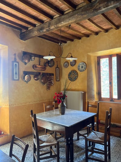 The image captures an interior space in Sinalunga, Italy. The room has a rustic aesthetic, characterized by exposed brick and wooden beams in the ceiling. The walls are painted in a warm, golden hue with decorative floral motifs. Several decorative plates and a collection of copper cookware are mounted on the wall. A table is positioned in the room, and chairs surround it. A vase of red flowers sits on the table. Natural light enters through a window. The floor appears to be tiled.
