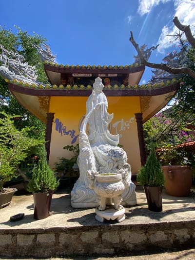 This is a full, angled shot of a large, white marble statue of a serene female figure, possibly a deity, with flowing robes and intricate details in the sculpture. She appears to be seated or reclined on a decorative base that resembles water or clouds, with a mythical creature, possibly a dragon or lion, carved into it. The statue is positioned outdoors, in what appears to be a temple courtyard or garden, given the presence of lush green trees and potted plants.
Behind the statue, there is a building with a traditional East Asian architectural style. The building features a yellow facade with blue calligraphy inscribed on it. The roof is adorned with decorative elements, including what look like golden spheres and stylized dragon-like figures.
The scene is bathed in bright sunlight, indicating it is daytime, likely mid-morning or afternoon. The sky is a clear, vibrant blue with a few scattered white clouds. The overall mood is peaceful and spiritual, consistent with a religious or cultural site.
The text in blue on the yellow wall appears to be in Vietnamese. The location is specified as Thôn 5, Vietnam. The presence of traditional architecture, the statue, and the Vietnamese text strongly suggest this is a scene from a Buddhist temple or a similar cultural landmark in Vietnam.