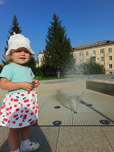 A young Caucasian girl, appearing to be around two years old, stands in a public square in Zlatoust, Russia, near a fountain on a sunny day. She is wearing a white lace hat, a light blue t-shirt, and a white skirt with a cherry pattern. The girl is holding a pinecone in her hands. The fountain is spraying water, creating a misty effect. In the background, there is a yellow building and several tall evergreen trees. The ground is paved with stone and metal grating. The sky is clear and blue.