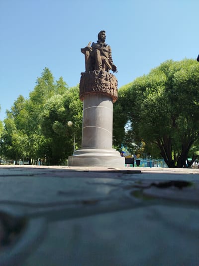 A low-angle, wide shot captures a bronze statue on a grey granite pedestal situated outdoors on a clear, sunny day. The statue depicts a seated man with his arms extended and an open book in his lap. The pedestal features an ornate carved band encircling its upper section. Behind the statue, a row of lush green trees forms a dense backdrop, with the bright blue sky visible above. In the middle ground, a lamp post and further trees are visible, along with glimpses of what appears to be a park or amusement area with colorful structures and fencing. A few indistinct figures are present in the background. The foreground is dominated by a textured, grey surface, possibly a paved area, with dark, wet-looking patches that reflect light. This statue is located in the city of Yalutorovsk, Russia.