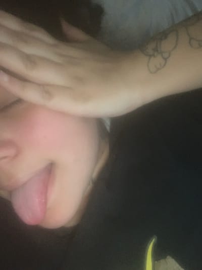 The image displays a close-up, rotated view of an individual's face and right arm. The specific location within the city of Lancaster, United States of America, is not discernible from the visual information provided. The individual's lower face, from the nose to the chin, is visible, characterized by a light skin tone and a flushed appearance on the cheek. The tongue is extended outwards from the mouth. The right hand covers the upper portion of the face, obscuring the eyes and forehead. A line-art tattoo, appearing to depict abstract or stylized cloud-like shapes, is visible on the individual's right forearm. The visible clothing is dark. The background is indistinct and dark, suggesting an indoor setting with low light or a blurred background. There are no other discernible subjects, objects, or interactions beyond the individual and their immediate actions.