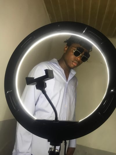 An indoor portrait displays a young male, estimated late teens to early twenties, with dark complexion and short, dark, textured hair. He wears a light-colored, open-collared long-sleeve shirt and square, gold-framed dark sunglasses. His mouth is slightly open, and he looks directly towards the viewer. A large, black, illuminated LED ring light is prominently positioned in the foreground, partially obscuring the lower half of the subject and framing his head and shoulders. A black smartphone holder, attached via a flexible gooseneck arm, extends from the ring light into the lower-left quadrant of the frame. The background consists of a plain, light beige wall, with a section of a light-colored ceiling, possibly paneled, visible in the upper-left corner. The specific location within Kirana, Nigeria, is not discernible from the visual evidence.