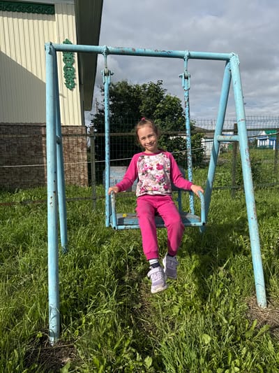 A young girl with brown hair, wearing a pink floral shirt and pink pants, lies on a blue metal structure in an outdoor setting. She is lying on her back, with her right arm bent and her hand resting on the metal. Her eyes are closed, and she appears to be relaxed or sleeping. The metal structure looks like a swing set or a part of a playground, painted light blue with some rust showing through.
The girl is positioned on a horizontal bar of the structure, with her legs extended. She is wearing grey sneakers with white laces and some black and white striped socks. The background consists of green grass and trees, suggesting a park or a rural area. The sky is overcast with grey clouds, indicating a cloudy day.
In the background, there are glimpses of buildings, possibly a house or sheds, with white and light yellow siding. There is also a metal fence and more green foliage. The overall scene is casual and relaxed, with the girl seemingly enjoying a moment of rest outdoors. The time of day is difficult to ascertain precisely due to the overcast sky, but it appears to be daytime. No visible text is present in the image. The location is specified as Bolshoy Turysh, Russia.