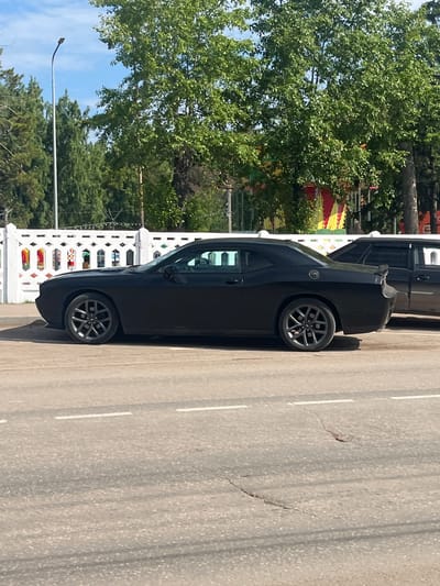 A black Dodge Challenger is parked on the side of a paved road in Oktyabrsky, Russia. The car is angled, with its driver's side facing the camera. A white decorative fence runs along the background, behind which are trees and some colorful structures that appear to be part of a fair or carnival. The sky is blue with some clouds, and a street light is visible on the left side of the frame. The road has visible lane markings and some cracks. Another black car is parked behind the Challenger. There are no people visible in the image.