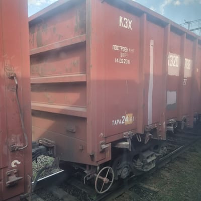 A red freight train car is parked on railway tracks. The car is marked with white lettering including "K3X", "ПОСТРОЕН КНР", "QRRS", "14.09.2011", and "TAPA 24.6T". Other markings and numbers are visible on the car and on adjacent cars. The ground around the tracks is covered with gravel and some vegetation. The sky is visible above the train car, with some clouds. This image was taken in the city of Kandyagash, Kazakhstan.