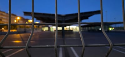The image depicts a wire fence in the foreground, with its metallic bars obscuring the background. Beyond the fence, at dusk or early night, is a blurred scene of an open area, possibly a parking lot or a public space. Several streetlights with a warm, yellow-orange glow are visible on the left side of the image, illuminating a building facade and casting reflections on the ground. In the middle ground, a large, dark, roof-like structure with an unusual, wing-like silhouette is prominent. Beneath this structure, there appear to be illuminated storefronts or buildings, with lights creating soft, bokeh effects. White lines, indicative of parking stalls or road markings, are faintly visible on the dark ground. The sky is a deep blue, transitioning to a darker hue at the top. The overall atmosphere is dim and somewhat indistinct due to the low light conditions and shallow depth of field. The specific location within Huesca, Spain, is not discernible from the visual elements present in the image.