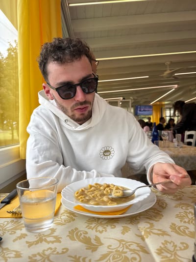 The image depicts an indoor dining scene at a restaurant in Anzola dell'Emilia, Italy. In the foreground, a male subject, approximately 20-30 years old, with short curly brown hair and facial stubble, is seated at a table. He is wearing black sunglasses and a white hooded sweatshirt adorned with a subtle gold or tan circular flower emblem on the left chest. His right hand holds a silver spoon, positioned above a white bowl containing ring-shaped pasta, consistent with tortellini, served in a light broth or sauce. The bowl rests on a white plate, which is placed on a folded orange napkin. To his left, a clear glass filled with water and a set of silverware (knife and fork) are visible.
The table is covered with a light cream-colored tablecloth featuring a repeating pattern of golden floral or scroll motifs. In the background, to the left, a large window with bright yellow curtains drawn to the side reveals outdoor greenery. The ceiling features exposed white wooden beams with multiple linear fluorescent light fixtures and a white ceiling fan. Further into the restaurant, other blurred patrons are visible at additional tables, indicating a communal dining environment. A blue bottle and a television screen displaying an image are faintly discernible in the distant background.