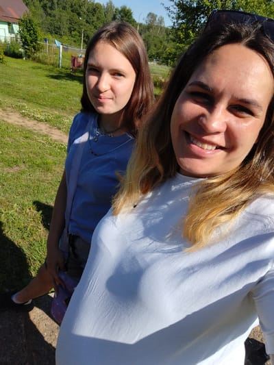Two young women are taking a selfie outdoors on a sunny day. The woman in the foreground is on the right, smiling broadly at the camera. She has long, blonde-highlighted hair and is wearing a white t-shirt. The woman behind her on the left is also looking at the camera with a slight smile. She has long, brown hair and is wearing a blue t-shirt and a light-colored shoulder bag with a patterned strap.
The background shows a grassy area with a few trees and a wooden fence. A small, light-colored building with a red roof is visible behind the fence, as are some clothes hanging on a clothesline and a red trash bin. The scene appears to be in a rural or suburban setting, possibly a dacha or a similar type of property. The bright sunlight and clear blue sky suggest it is daytime, likely late morning or early afternoon. The overall mood seems relaxed and happy. There is no visible text in the image.