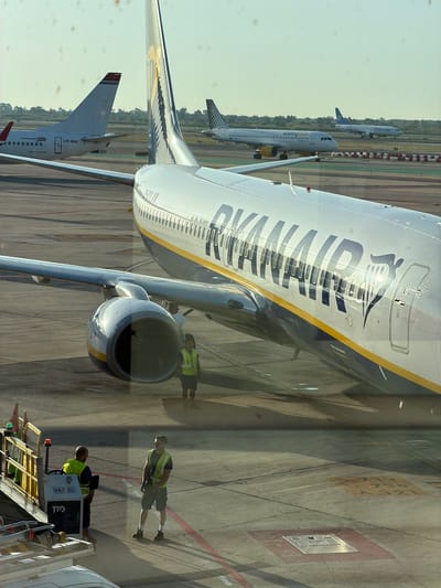 This media file captures a scene at El Prat de Llobregat, Spain, likely an airport tarmac. The main subjects are several commercial airplanes and a few individuals wearing high-visibility vests.
In the foreground, a large Ryanair airplane dominates the frame, its distinctive blue and yellow livery visible. Part of its engine and fuselage are clearly depicted. Behind it, another airplane, possibly a Vueling, is also visible, with "vueling.com" printed on its side. Further in the background, a third airplane, identified as "Alteuropa," is present.
The scene depicts ground operations at an airport. The individuals in high-visibility vests appear to be airport personnel engaged in their duties, possibly servicing the aircraft or directing ground traffic. One person is standing near the Ryanair plane, while another is seated on a vehicle with "TID" and "RYR-E-011" visible on its side.
The lighting suggests it is daytime, likely with sunny weather, as indicated by the bright illumination and the shadows cast by the planes and people. The ground is paved with asphalt, marked with lines and some colored squares, typical of an airport environment. The overall impression is one of an active airport during daylight hours, with commercial aviation activities underway.