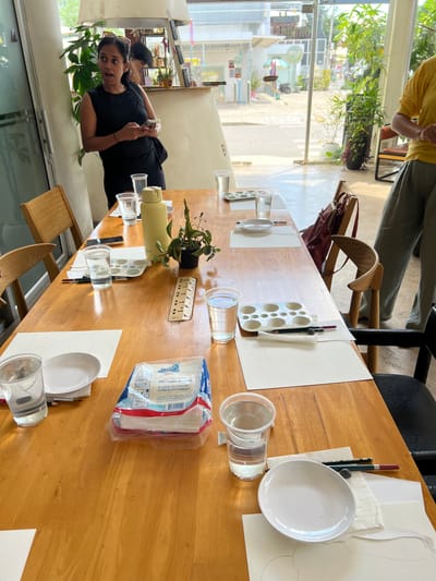 In a Siem Reap art studio, a woman with dark hair tied back, wearing a black sleeveless shirt and leggings, stands at a wooden table, holding a smartphone and looking off-camera with an open-mouthed expression. She appears to be interacting with someone or observing something outside the frame. The table is set up for an art class, with several place settings including white paper with outlines of objects, small white plates, glasses of water, and paint palettes. A yellow water bottle, a small potted plant, a ruler, and a package of tissues are also on the table. Sunlight streams in from large windows, illuminating the space and revealing a street scene with buildings and greenery outside. The overall atmosphere suggests a creative and educational environment.