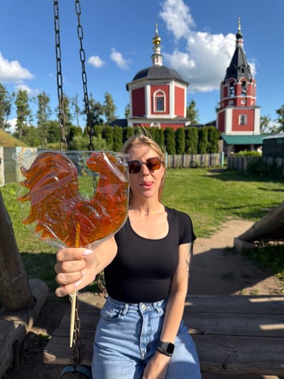 A woman is sitting on a wooden swing in Suzdal, Russia, holding a large, rooster-shaped lollipop. The lollipop is made of translucent amber-colored candy and is wrapped in plastic. The woman is wearing a black t-shirt, blue jeans, and sunglasses. She is sticking her tongue out playfully. In the background, there are two red and white Russian Orthodox churches with golden domes and a black spire. The churches are surrounded by green trees and a wooden fence. The sky is blue with some white clouds. The scene is set in a park or historical area.