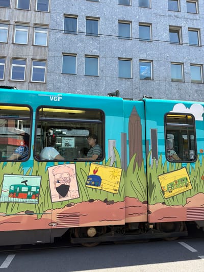 A teal-colored tram decorated with cartoon drawings is parked next to a modern building. The building, likely an office or apartment complex, has a gray, speckled facade with numerous uniformly spaced rectangular windows. The tram is adorned with bright, colorful artwork depicting a grassy landscape, a cityscape with a tall skyscraper, and various cartoon characters on yellow rectangular cards.
Inside the tram's windows, several people can be seen. In the upper window, a person with glasses wearing a striped shirt is visible, seemingly looking out. In the lower window, another person is visible, wearing a floral-patterned shirt. The exterior of the tram features the "VGF" logo prominently. Some of the cartoon drawings on the tram are children's drawings, with one depicting a sun and stick figures, and another a stylized anime character. The scene suggests a sunny day, given the bright lighting and shadows. The presence of the "VGF" logo indicates the tram is likely in Frankfurt, Germany, and in service for the local public transport authority. The overall impression is one of a vibrant urban environment with public art integrated into everyday infrastructure.