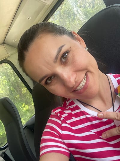 A woman with light skin and brown eyes is smiling at the camera. She is wearing a red and white striped shirt and a black necklace. She appears to be inside a vehicle, possibly a bus or van, based on the presence of a black seat and a window showing green trees. The numbers "21" and "22" are visible on a white panel near the window. Although I can't verify that the location is Donskoe, Russia from the image alone, the presence of the numbers suggests that the scene is from a transit system. The woman has an earring in her left ear. She is holding up her index finger.