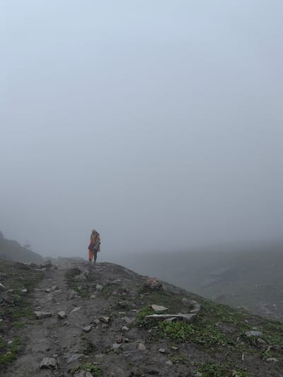 A lone person, wearing orange and brown clothing, stands on a rocky, dirt path in Dhundi, India. The person is facing to the left and is carrying a backpack. The environment is hazy with fog, obscuring the background which appears to be a mountainous landscape. Rocks and sparse green vegetation are visible on the path and the surrounding slope. The overall atmosphere is subdued due to the fog and muted color palette.