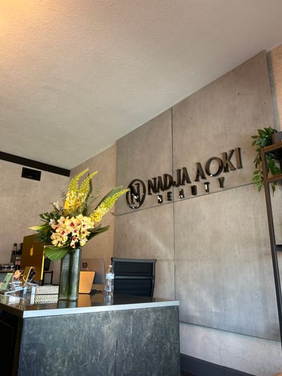The media file shows a retail space with a concrete ceiling and textured walls. A prominent feature is the black, three-dimensional lettering on the ceiling, which reads "NADJA AOKI BEAUTY".
In the foreground, there is a large floral arrangement with yellow orchids and other greenery. The arrangement is displayed in a golden planter or vase. Behind the flowers, a counter area is visible, featuring what appears to be a laptop, a water bottle, and some other electronic devices. The counter itself has a dark, textured surface.
The scene appears to be an interior, possibly a salon or boutique, given the "BEAUTY" signage and the stylish presentation. The lighting suggests it is daytime, with natural light entering from an unseen source, casting a warm glow on the textured wall. The overall aesthetic is modern and minimalist, with a focus on natural textures and materials. There are no people visible in the image. The location is identified as Deerfield Beach, United States of America, though this context is not visually evident in the image itself.