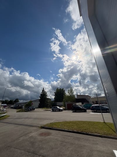 The image depicts an outdoor scene on a sunny day with scattered clouds. A modern industrial building with a corrugated metal facade occupies the right side of the frame, with the sun creating lens flare and reflections on its surface. The foreground is an asphalt road and a patch of green grass. In the background, several cars are parked. Beyond the parking area, there are industrial buildings, trees, and a prominent evergreen tree. The sky is a vibrant blue with bright white and dark grey clouds, indicating dynamic weather. The location appears to be in an industrial area, possibly on the outskirts of Sauleskalns, Latvia, given the architectural style and the presence of industrial facilities. There are no people visible in the image.