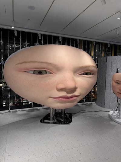 A close-up, eye-level shot showcases a large, hyperrealistic mannequin head with a round, smooth forehead and visible freckles. The mannequin's left eye, facing the camera, is a detailed iris of green and brown hues, with a slightly downturned gaze. The right eye is partially visible and also appears to be looking slightly down. The lips are a soft pink, with a subtle, closed-mouth expression. The mannequin is mounted on a metal stand, with another smaller mannequin head partially visible in the background to the right. The environment appears to be an indoor exhibition space, with a speckled grey floor and a backdrop of decorative metallic hanging elements and a wall panel. The lighting is overhead, with spotlights visible. The scene is located in the retail and exhibition district of Chuo, Japan.