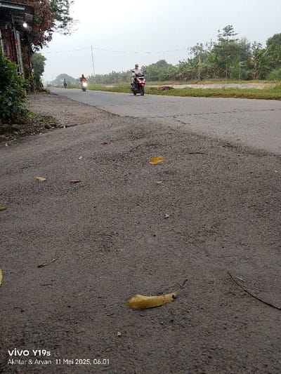 The image shows a close-up view of an asphalt road in Kecamatan Haurgeulis, Indonesia, located at latitude -6.42 and longitude 107.977. The road is empty except for three motorbikes traveling in the distance. The road is paved with asphalt and has a rough surface. There are a few fallen leaves scattered on the road. The image is taken from a low angle, looking down the road. The background is blurred, indicating a hazy or overcast sky. The photo was taken on May 11, 2023, at 6:01 AM.