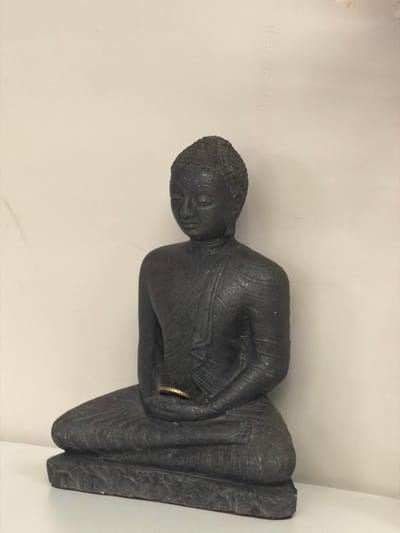 A dark grey, textured Buddha statue is seated in a meditative pose on a white surface against a light beige wall. The statue is small enough to fit within the frame, with its hands resting on its lap, one hand holding a thin, gold ring. The statue's face is downcast, and its robes are depicted with textured lines. The lighting is soft, casting minimal shadows. The location is not specified in the image or its context.