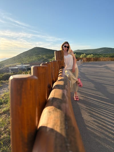 A young woman is standing next to a wooden fence on a paved path with a scenic mountainous landscape in the background. The woman has blonde hair and is wearing sunglasses, a white tank top, and beige pants. She is leaning against the fence with one leg crossed over the other, wearing pink sandals. The sun is low in the sky, casting long shadows across the path. In the distance, there are hills covered in greenery, a few buildings, and a clear blue sky with wispy clouds. The location is identified as Krimovice, Montenegro.