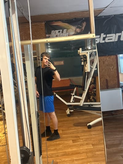 The image is taken indoors, presumably in a gym, evidenced by the presence of various exercise machines. A man is visible in the foreground, seemingly holding a phone to his ear, possibly on a call. He is wearing a black t-shirt, blue shorts, black socks, and black shoes. A banner with the word "Start" is visible in the background. The location is in the city of Elhovo, Bulgaria, as indicated by the Cyrillic text on the banner. The gym appears to have wooden floors and walls.