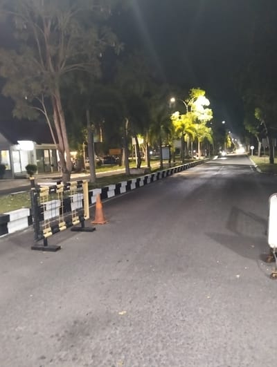 The image captures a nighttime scene on an asphalt road bordered by trees and a white and black striped barrier. In the foreground, a black and yellow striped barricade stands on the left, accompanied by an orange traffic cone. Further down the road, palm trees and other foliage line the sides, illuminated by streetlights. The road stretches into the distance, curving slightly to the right, with more streetlights visible along its path. On the left side of the road, a building with windows and lights can be seen. The overall setting appears to be an outdoor area, possibly a campus or a park, in Tambontunong, Indonesia, as indicated by the location context. The time of day is clearly nighttime, indicated by the dark sky and artificial lighting. No people are visible in the frame. The image conveys a sense of quietness and emptiness due to the absence of activity and people. No visible text is present in the image.