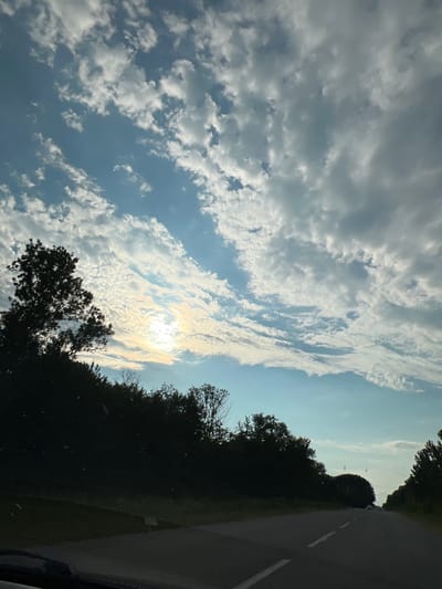 The image depicts a view from a vehicle traveling on a road, likely in or near Ihrovytsia, Ukraine. The sky is filled with a dramatic display of clouds, ranging from wispy, textured formations to larger, more solid masses. The sun is partially obscured by these clouds, casting a bright, diffused light that creates a slight rainbow effect in the upper atmosphere. Silhouetted against the sky on the left side of the frame is a dense line of trees and foliage that borders the road. The road itself is paved, with white lane markings visible. The overall impression is one of a natural landscape under a dynamic sky, observed from the perspective of someone in transit.