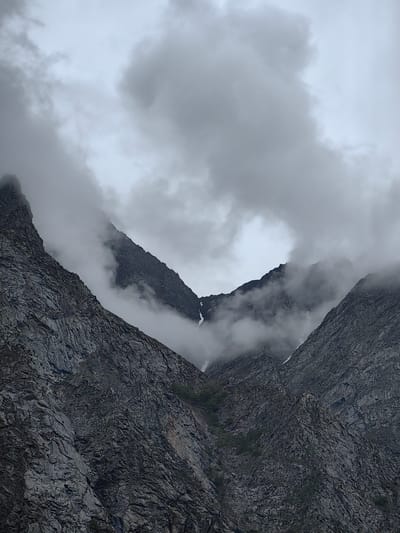 The image depicts a dramatic mountain landscape in Jispa, India, under a heavy, overcast sky. Two massive, rugged rock faces dominate the frame, rising steeply towards the sky and converging in the center, forming a V-shaped valley. The mountains are textured with intricate patterns of cracks, fissures, and rocky protrusions, suggesting their immense age and the forces of erosion that have shaped them.
Wisps and thicker banks of dense, grey clouds cling to the upper reaches of the mountains, obscuring some of their peaks and adding a sense of mystery and foreboding to the scene. In the central valley, more clouds swirl and drift, partially veiling the deeper recesses of the mountain range. There are hints of sparse, green vegetation clinging to the lower slopes of the rocks, a testament to life's tenacity even in such harsh environments. Patches of snow are visible on the higher elevations, indicating a cold climate. The overall impression is one of raw, untamed natural power and the immense scale of the Himalayas.