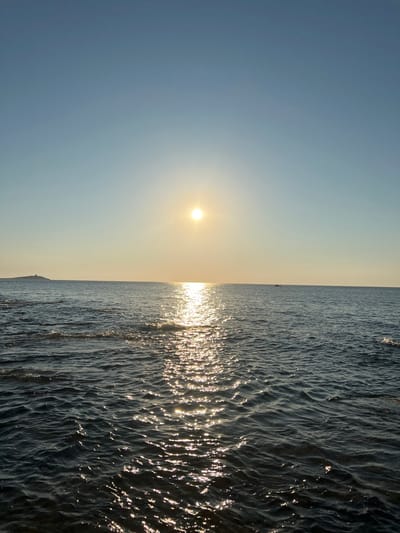 The image depicts a serene seascape with the sun low in the sky, casting a warm glow. The sun, a bright orb, is positioned in the left half of the frame, its light reflecting brilliantly off the rippling surface of the dark blue water, creating a shimmering path towards the horizon. The water occupies the right two-thirds of the image, showing the texture of small waves.
In the far distance, a very faint silhouette of what appears to be a small boat or a distant landmass can be seen on the horizon. The sky is a soft gradient of pale blue and warm yellow, indicating either sunrise or sunset. The overall atmosphere is tranquil and peaceful, suggesting a quiet moment of observation by the sea. The scene is likely a coastal location, and given the provided context of Palermo, Italy, it could be a view of the Mediterranean Sea. No text is visible in the image. The orientation of the image is vertical.