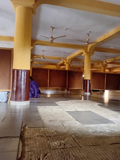 The interior showcases rows of wooden pews under a high ceiling supported by yellow-painted beams. Several ceiling fans are evenly spaced along the ceiling. The floor features a mix of smooth tiles and a rougher, potentially unfinished section near the wall. A purple jacket lies on one of the pews. The perspective gives a somewhat distorted view, emphasizing the height of the room and the repetition of the architectural elements.