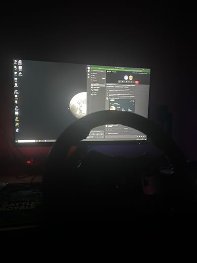 The image shows a dimly lit room with a computer monitor as the central focus. The monitor displays a desktop with various icons on the left side, including "This PC," "Recycle Bin," and applications like "Epic Games" and "Steam." The background of the desktop is a dark image of the moon. On the right side of the monitor, a Discord-like application interface is visible, showing user profiles, chat messages, and channel lists. The chat messages appear to be in Bulgarian. In the foreground, a dark silhouette of a gaming steering wheel is present, obscuring the lower part of the monitor and the desk. The ambient lighting suggests a dark environment, possibly a gaming setup. There are no visible people. The image does not contain any information about the city of Madara, Bulgaria.