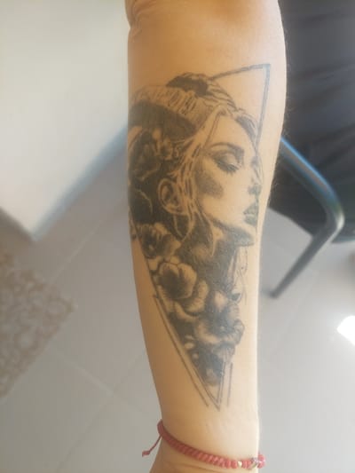 The image shows a close-up of a person's forearm with a black and grey tattoo of a woman's face and flowers within a triangle. The tattoo is detailed and appears to be in progress or slightly faded. A thin red braided bracelet with small beads is visible around the wrist. The background is blurred and out of focus, suggesting an indoor environment with light sources creating shadows. The overall impression is a personal portrait tattoo on a person's arm.