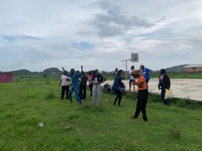 A group of young people, appearing to be in their late teens or early twenties, are gathered on a grassy field with a basketball court in the background. The sky is overcast with dramatic clouds, suggesting it might be late afternoon or early evening. The overall atmosphere appears energetic and joyful, as many of the individuals have their arms raised and are in motion, as if dancing or celebrating.
There are at least ten people visible, dressed in casual clothing. Some are wearing t-shirts and pants, while others are in more vibrant, patterned outfits. One person in the foreground is wearing an orange t-shirt and black pants, and another is in a blue and red patterned outfit. There's a pink building visible on the far left of the image, and a more modern brick building in the distance on the right, possibly indicating a school or community center. The landscape includes rolling hills in the background.
The activity seems to be informal, with the group enjoying themselves outdoors. The presence of a basketball hoop and court suggests a potential link to sports or recreational activities, but the current focus seems to be on a spontaneous group dance or energetic interaction. The ground is covered in green grass with some patches of dry weeds and scattered white flowers. A crumpled plastic bag is visible on the grass in the foreground. The location is identified as Gwafan, Nigeria.