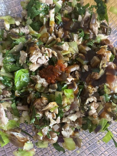 A close-up image displays a prepared food dish, likely a mixed salad or a savory stir-fry, presented in a clear, textured bowl. The dish is a vibrant combination of various ingredients. Prominently featured are small, irregular chunks of light-colored meat, possibly chicken or pork, which are mixed with abundant green leafy vegetables, appearing to be a mix of lettuce or cabbage and fresh herbs like cilantro or parsley. Darker, softened vegetable pieces, potentially cooked onions or mushrooms, are also visible, adding to the texture and color palette.

A glossy, dark brown sauce or dressing is generously drizzled over the entire mixture, creating a rich sheen. Near the center, a distinct blob of reddish-orange condiment, possibly a chili paste, salsa, or a spicy relish, stands out, suggesting a potential kick of flavor. The ingredients are finely chopped and appear to be thoroughly combined. The container itself is transparent, with a visible grid-like pattern on its surface, indicating it might be made of plastic or glass.

The image provides no cues regarding the time of day, weather, or any emotions. No text is visible within the frame. The provided location context for this media file is Egkomi Lefkosias, Cyprus, although this information is not visually represented in the image itself.
