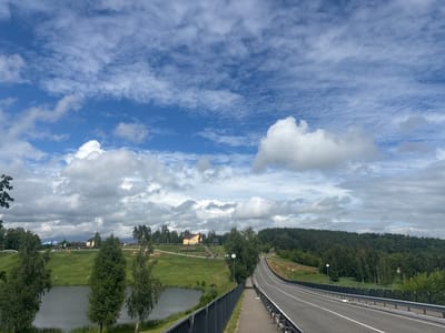 The image shows a road leading uphill through a rural landscape under a partly cloudy sky. On the left side of the road, there is a body of water and a grassy slope with trees and buildings on top. The buildings appear to be houses or a complex of some sort, with one prominent yellow building. There are also some decorative elements and signage on the slope. The right side of the road is bordered by a fence and then a dense forest. The road itself is asphalt with white lane markings and appears to be a two-lane highway. The overall scene suggests a scenic route in a countryside area. This location is identified as the Капыльскі сельскі Савет, Belarus, likely a point of interest or a route through the region.