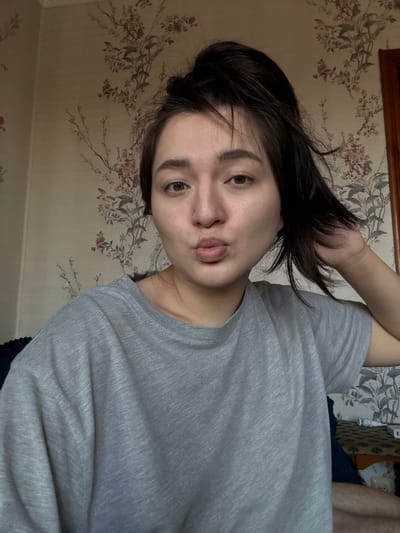 A young adult female with short, dark hair is angled towards the camera. She is wearing a gray t-shirt. Her hand is raised near her head. The background is a wall with a floral pattern. The location is potentially a domestic interior in Kurovskoye, Russia, if that information is presumed from context.