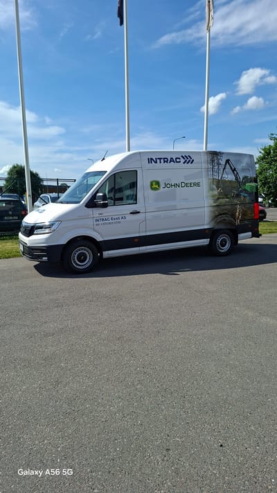 In the city of Loo, Estonia, under a bright blue sky with scattered clouds, a white panel van is parked on an asphalt surface. The van is branded with "INTRAC" and "JOHN DEERE" logos. On the side of the van, there is text that reads "INTRAC Eesti AS tel: +372 603 5700". The van also features a large graphic image on its rear half depicting a forest scene with a green John Deere excavator. In the background, two flagpoles are visible, one with a dark flag, and a fence with some trees beyond it. The overall scene is one of an outdoor industrial or commercial area on a sunny day.