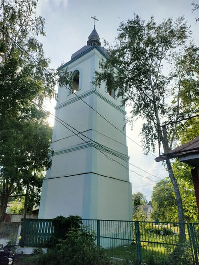 The image depicts a tall, pale blue and white bell tower with a grey, octagonal roof topped by a cross. The tower has arched openings on its upper level. It is surrounded by lush green trees, and overhead power lines crisscross the sky. In the foreground, there is a green metal fence, and behind it, overgrown vegetation and parts of buildings. To the right, a portion of a reddish-brown roof is visible. The sun is visible through the trees, creating a bright glare. The overall scene suggests a rural or suburban setting in Nosivka, Ukraine, likely a church or religious building.