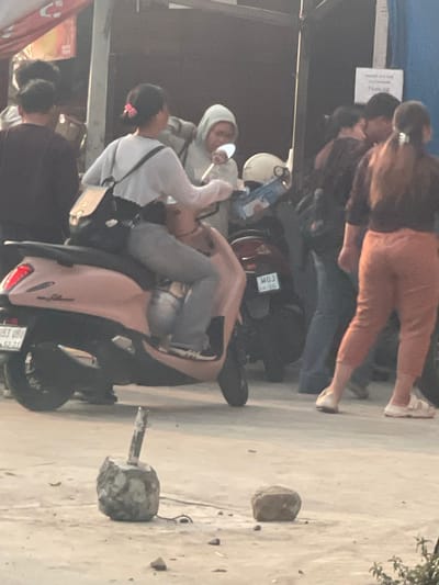In the city of Tangerang, Indonesia, a street scene unfolds with several individuals interacting around motorbikes. In the foreground, a light pink scooter is prominent, with a young woman wearing a white long-sleeved top, grey pants, and sneakers seated on it. She has a backpack on her shoulders and appears to be holding a white object. Another scooter, dark in color, is parked nearby. Behind the seated woman, another person wearing a light grey hooded jacket is visible. In the background, a group of people are gathered, some partially obscured. The environment suggests an urban setting, possibly a market or roadside, with elements like a stall canopy visible on the left and a sign on a wall in the middle ground. The ground is paved with what appears to be concrete or asphalt, with a few scattered stones and a concrete object with a metal rod sticking out of it in the lower left foreground. The lighting is soft, indicating it might be early morning or late afternoon.