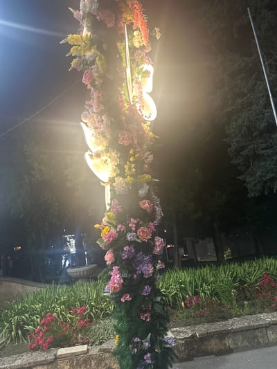The image shows a lamppost decorated with artificial flowers at night. The lamppost is adorned with a cascade of colorful flowers in shades of pink, purple, yellow, and white, intertwined with greenery. Two light fixtures are visible on the lamppost, emitting a warm glow that illuminates the flowers and creates a soft ambiance. In the background, trees and a stone wall are discernible, suggesting an outdoor urban setting. A fountain and some distant lights are also visible, hinting at a public space within the city. The overall impression is one of festive decoration, likely intended for an event or a seasonal display. The image is taken from a low angle, emphasizing the height and floral embellishments of the lamppost. The location is specified as Shumen, Bulgaria.