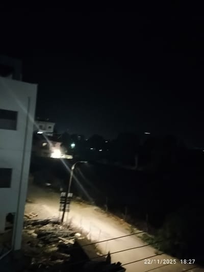 This nighttime image presents an elevated view of a semi-urban or suburban area. The scene is dominated by darkness, with artificial lights providing the main illumination. The timestamp "22/11/2025 18:27" is visible in the bottom right corner, indicating the date and time of the photograph.
On the left side of the frame, a light-colored, multi-story building with dark windows is partially visible, suggesting the vantage point. Below this building, along what appears to be a dirt or unpaved road, there is a pile of materials, including what looks like rebar and construction debris, brightly illuminated.
A tall streetlamp stands beside the road in the middle ground, emitting a strong light that casts an elongated glow across the path and creates noticeable lens flares. The unpaved road extends into the distance, bordered by fences or low structures on its right side.
The background is mostly dark, with indistinct silhouettes of trees or distant landforms. Several small, distant lights are scattered across the dark horizon, possibly from other buildings or vehicles. A thin, bright horizontal light streak is also visible in the upper right portion of the image, potentially an aircraft or a distant light source.
No human activity or individuals are visible within the frame, contributing to a quiet and somewhat desolate atmosphere. The overall impression is one of an evening landscape in North Nicosia, Northern Cyprus, characterized by the interplay of artificial light and deep shadows.