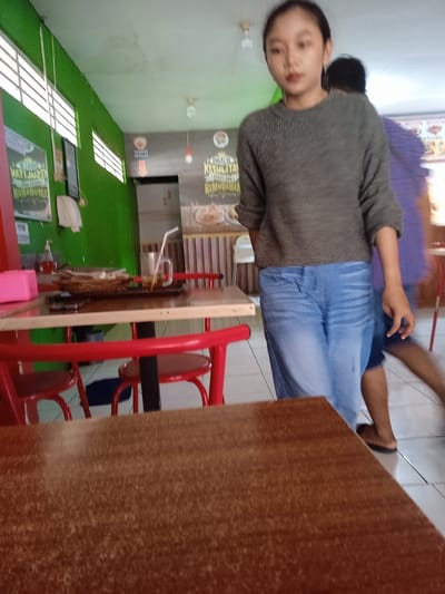 The image depicts an indoor scene, likely a cafe or restaurant, in Kaliwungu, Indonesia. In the foreground, a portion of a brown wooden table is visible. Behind it, a young woman with dark hair is standing, wearing a grey knitted sweater and blue jeans. She is facing towards the camera with a neutral expression. To her right and slightly behind her, another person in a purple shirt is visible, with their back to the camera, and their face blurred. In the mid-ground, a red-framed table with red chairs is set up. On the table, there is a brown tray with a glass of iced tea and a plate of food. In the background, a green wall with horizontal window slats and promotional posters is present. The overall lighting suggests an indoor environment with overhead lights.