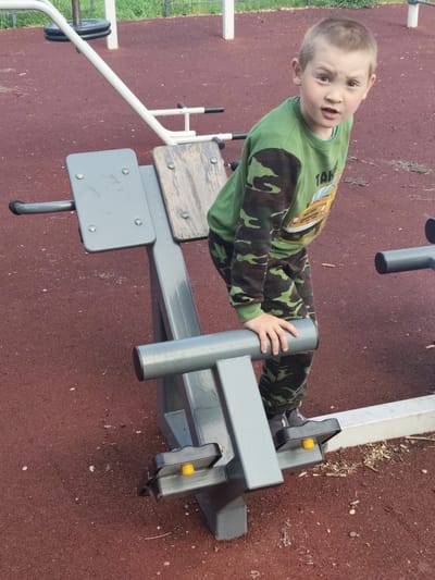 A young boy with light brown hair, wearing a camouflage-patterned tracksuit and a green long-sleeved shirt with a yellow car graphic, stands on an outdoor exercise machine. The boy is looking towards the camera with his mouth slightly open. The exercise machine is made of grey metal with a wooden seat and grey padded grips. It has yellow footplates at the bottom. The ground around the machine is a reddish-brown, soft surface, likely rubberized playground material, with scattered small white pebbles and twigs. In the background, a white metal frame of another piece of playground equipment is visible, along with green grass and what appears to be a fence. This scene is likely located in a public park or playground in Askiz, Russia, given the context.
