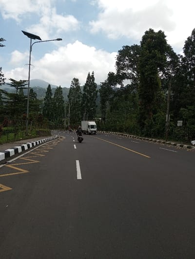 The scene displays a two-lane asphalt road in Boommlog, Bhutan, under a partly cloudy sky. On the left, a curb with black and white striped markings runs alongside yellow zigzag road markings painted on the asphalt. A tall street light pole, featuring a solar panel and integrated light fixture, stands on the left verge. A motorcyclist, wearing dark clothing, operates a dark motorcycle in the left lane, heading away from the viewer. Further ahead in the right lane, a white box truck is also moving away from the viewer. A smaller vehicle or motorcycle is partially visible behind the truck in the distance. The road is marked with a solid white center line, a broken white line, and a solid yellow line on the right. The environment is characterized by dense vegetation, including numerous coniferous and broadleaf trees lining both sides of the road and covering distant mountainous terrain. Utility poles are visible among the trees.
