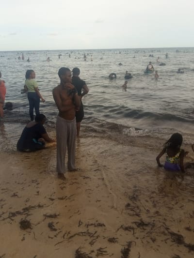 This image depicts a densely populated beachfront scene in Shanzu ward, Kenya, under an overcast sky. The foreground features damp, light-brown sand littered with dark organic debris, meeting shallow, turbid water with small breaking waves.
In the central foreground, a shirtless young male, wearing grey sweatpants, stands in ankle-deep water. His right arm is bent across his chest. To his immediate left, a female in a light green top and dark trousers stands partially submerged in the water. Behind the central male, a second male in a dark t-shirt and cap is partially visible, facing away from the camera. In the lower left quadrant, a female in dark attire crouches at the water's edge. In the lower right, a child with dark hair, wearing a multi-colored swimsuit, sits in the shallowest water.
The midground and background are dominated by a large body of water (ocean or large lake) teeming with an estimated 50-100 individuals of various ages. These people are engaged in swimming and wading. Several black inner tubes are clearly visible among the swimmers, utilized as flotation devices. The water in the mid-distance appears darker and more uniform. The horizon line where the water meets the sky is indistinct against the uniformly pale, overcast sky, indicating subdued lighting conditions.