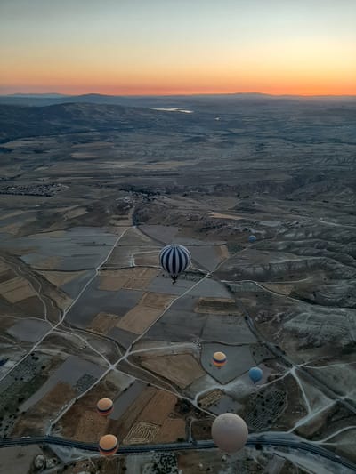 This aerial photograph captures the dawn sky over Avanos, Turkey, with hot air balloons dotting the landscape. The sky transitions from a pale blue at the top to a vibrant orange and pink near the horizon, illuminating rolling hills and valleys. The terrain below is a patchwork of agricultural fields, some fallow and others cultivated, divided by dirt roads and pathways. A winding highway runs through the lower portion of the frame. Several hot air balloons, varying in size and color, are visible: one large balloon with blue and white stripes dominates the center-left of the image, while others in shades of yellow, blue, and beige are dispersed across the scene. The balloons are floating at different altitudes, some appearing quite close to the ground and others higher in the sky. The overall impression is one of a serene and picturesque sunrise over a rural landscape.